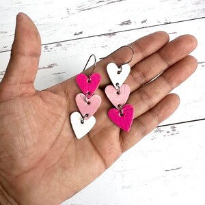 May include: A pair of dangle earrings with three layered heart charms. The hearts are white, light pink, and hot pink.