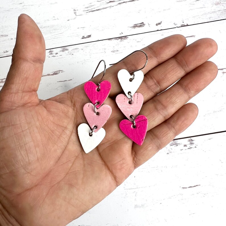 May include: A pair of dangle earrings with three layered heart charms. The hearts are white, light pink, and hot pink.
