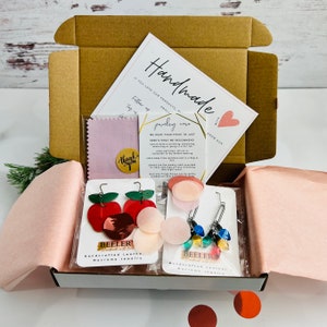 May include: A brown cardboard box with a pink and white tissue paper lining. The box contains three pairs of handmade macrame earrings. The earrings are made of leather and feature a variety of colors and designs. The box also includes a small pink cleaning cloth and a card with the text "Handmade Jewelry Care".
