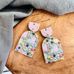 May include: A pair of dangle earrings with a floral and ghost pattern. The earrings are made of polymer clay and have a cork backing. The earrings are pink and green.