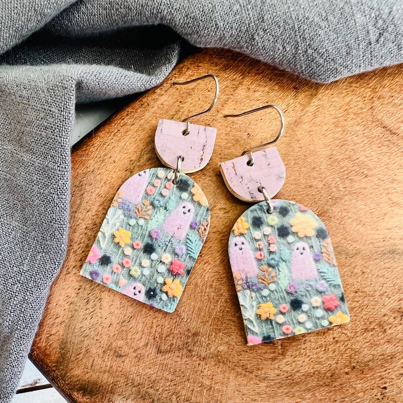 May include: A pair of dangle earrings with a floral and ghost pattern. The earrings are made of polymer clay and have a cork backing. The earrings are pink and green.