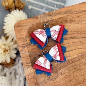May include: A pair of baseball-themed earrings. Each earring features a bow shape with a white baseball design, red accents, and blue backing. The earrings are attached to silver-colored ear wires.