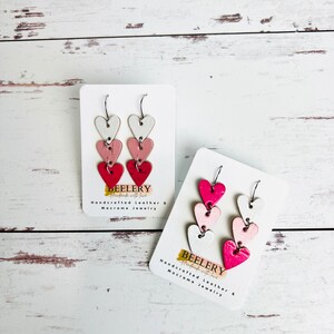 May include: A pair of dangle earrings with three layered hearts. The hearts are white, pink, and red. The earrings are on a white card with the text "BEELERY Handcrafted Leather & Macrame Jewelry".
