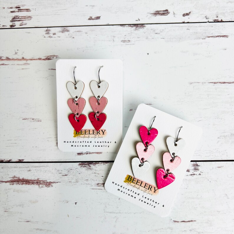May include: A pair of dangle earrings with three layered hearts. The hearts are white, pink, and red. The earrings are on a white card with the text "BEELERY Handcrafted Leather & Macrame Jewelry".