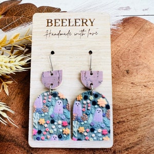 May include: A pair of purple and green ghost earrings with a floral pattern. The earrings are made of cork and have a silver hook. The earrings are displayed on a wooden card with the text "BEELERY Handmade with love".