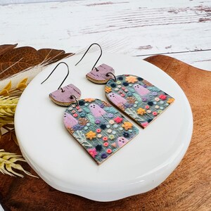 May include: A pair of dangle earrings with a whimsical design featuring ghosts and flowers on a purple and green background. The earrings are made of cork and have a silver hook.