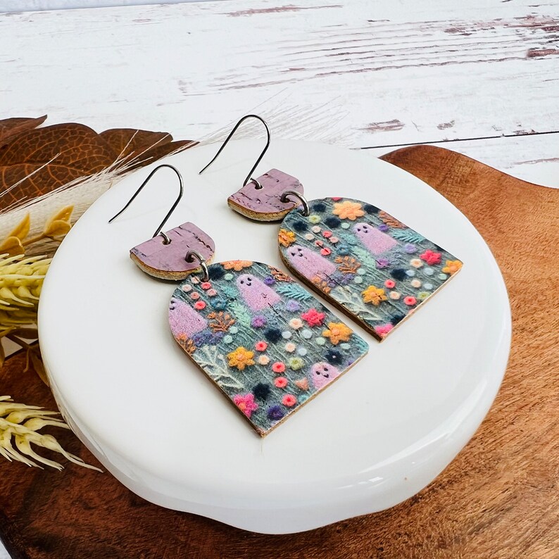 May include: A pair of dangle earrings with a whimsical design featuring ghosts and flowers on a purple and green background. The earrings are made of cork and have a silver hook.