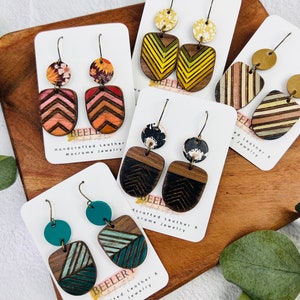May include: A collection of handcrafted leather and macrame jewelry earrings. The earrings feature various geometric designs and colors, including chevron patterns, stripes, and floral accents. The earrings are displayed on white cards against a wooden background.