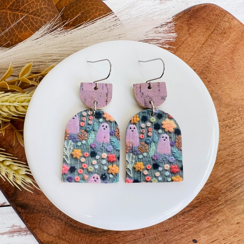 May include: A pair of dangle earrings with a purple and brown cork base and a top piece featuring a whimsical ghost pattern with flowers and leaves on a dark blue background.