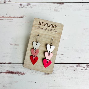 May include: A pair of dangle earrings with three layered hearts. The hearts are white, pink, and red. The earrings are on a wooden display card with the text "BEELERY Handmade with love".