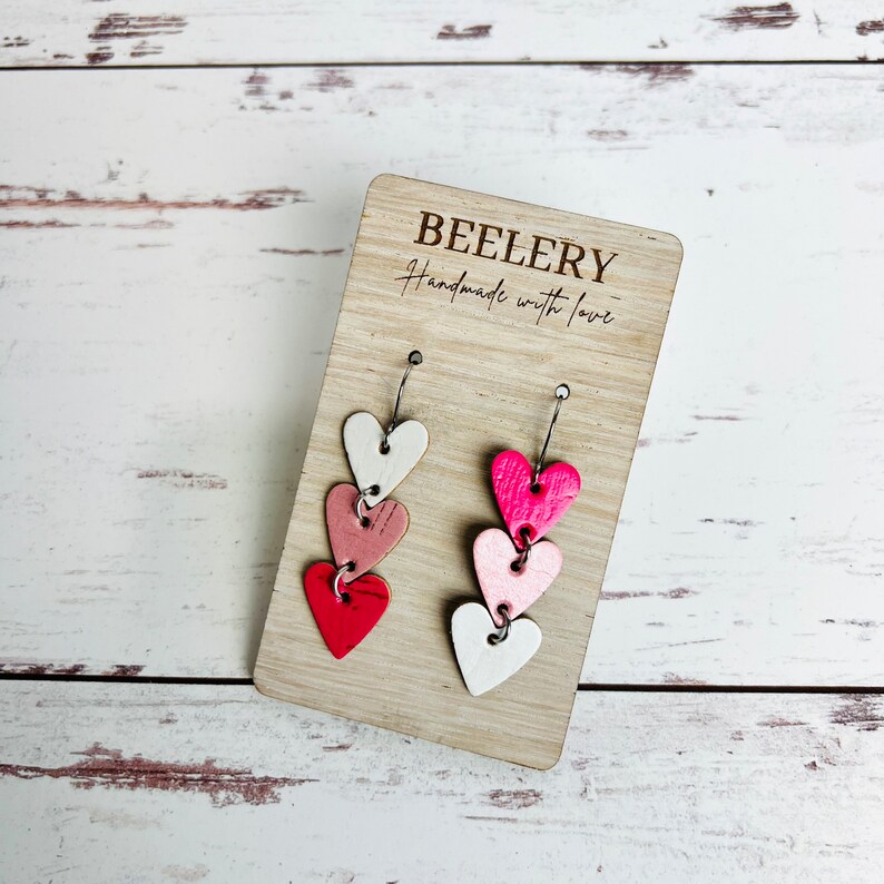 May include: A pair of dangle earrings with three layered heart charms. The hearts are white, pink, and red. The earrings are displayed on a wooden card with the text "BEELERY handmade with love".