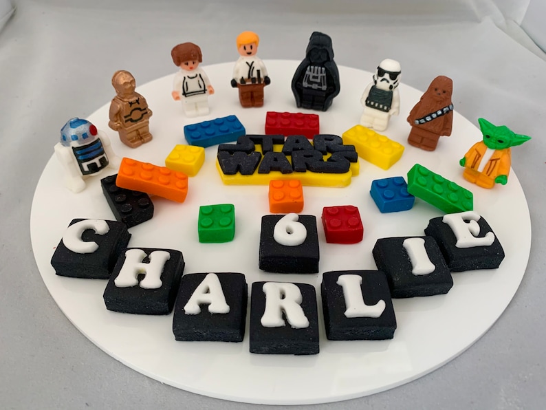 A Star Wars inspired cake topper set which includes Luke, Lea, R2D2, Yoda, Darth Vader, Chewbacca, Stomp Trooper, Star Wars Logo, 12 Lego blocks, Name and age on square tiles