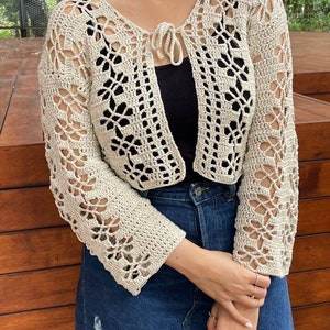 May include: A beige crochet cardigan with a tie at the neckline. The cardigan has a delicate openwork pattern and long sleeves.