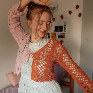 May include: A woman wearing a crocheted cardigan with a pink and orange color scheme. The cardigan has a tie at the front and is made with a lacy stitch.