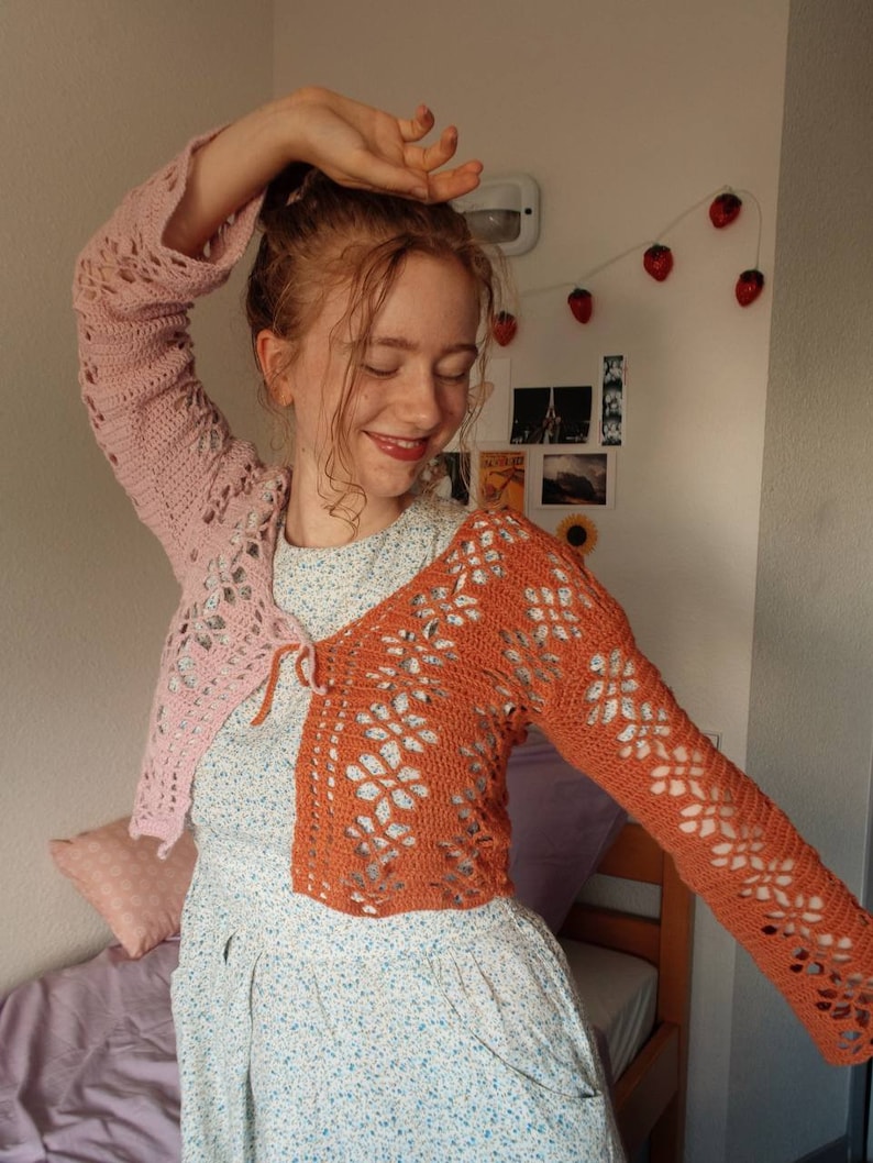 May include: A woman wearing a crocheted cardigan with a pink and orange color scheme. The cardigan has a tie at the front and is made with a lacy stitch.