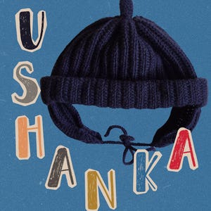 May include: Navy blue Ushanka hat with ear flaps and a top knot. The hat has the word "USHANKA" in colorful, blocky letters on a blue background. The hat is made of a ribbed knit material.