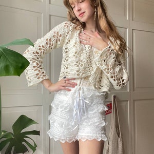 May include: A white crocheted cardigan with a delicate floral pattern, worn over a white lace skirt with a ruffled hem.