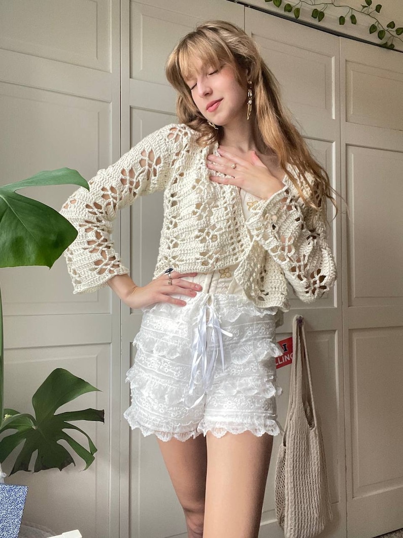 May include: A white crocheted cardigan with a delicate floral pattern, worn over a white lace skirt with a ruffled hem.