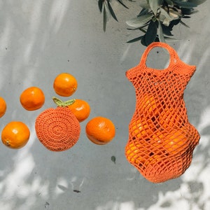 May include: A crocheted orange mesh bag filled with oranges. The bag is hanging from a white surface with a few oranges scattered around it.