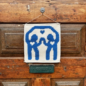 Puede incluir: Un tapiz de pared de crochet azul y blanco con un diseño geométrico. El tapiz está sujeto a una puerta de madera con un gancho de metal.