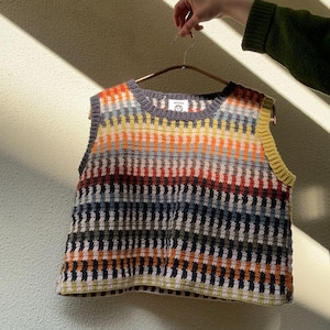 Può includere: Un top senza maniche all'uncinetto colorato con un motivo a righe nei toni dell'arancione, giallo, blu e grigio. Il top è tenuto da una gruccia. Il capo ha uno scollo tondo e un aspetto strutturato.