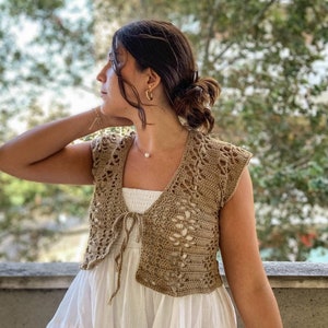May include: A brown crocheted sleeveless top with a tie at the front. The top is worn over a white dress.