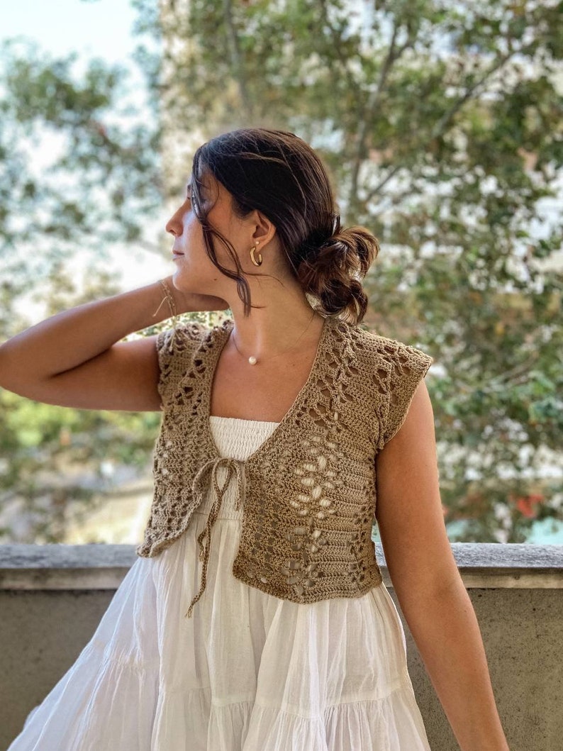 May include: A brown crocheted sleeveless top with a tie at the front. The top is worn over a white dress.