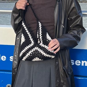 Peut inclure: Une personne portant un imperméable noir, un pull à col roulé marron et un sac en crochet noir et blanc. La personne porte une jupe plissée grise.