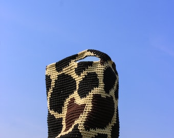 May include: A black and tan crocheted tote bag with a giraffe print pattern. The bag is sitting on top of a wooden post.