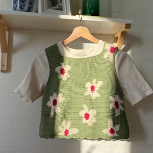 Puede incluir: Un top verde sin mangas de ganchillo con detalles de flores blancas y rojas. El top tiene un dobladillo festoneado y está colgado de una percha de madera.