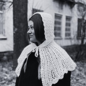 Puede incluir: Una capucha y una capelina de ganchillo blancas con un borde festoneado. El diseño calado de la capelina es visible. La capucha está atada al cuello con un cordón blanco. La persona lleva un abrigo negro.
