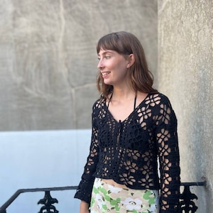 May include: A black crochet top with a V-neck and long sleeves. The top has a delicate openwork pattern and is worn with a floral print skirt.