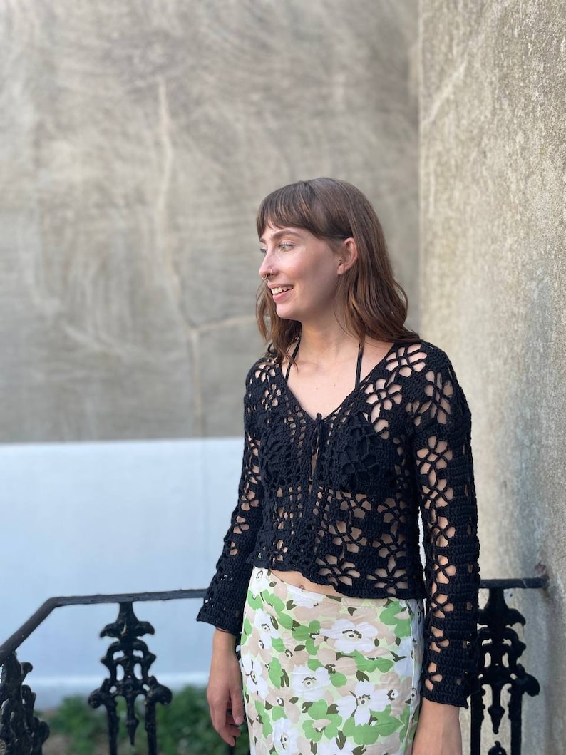 May include: A black crochet top with a V-neck and long sleeves. The top has a delicate openwork pattern and is worn with a floral print skirt.
