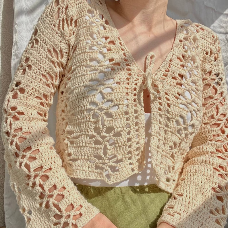May include: A cream-colored crocheted cardigan with a tie front and long sleeves. The cardigan has a delicate openwork pattern.