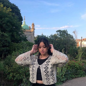 May include: A woman wearing a white crocheted cardigan with a tie front and bell sleeves. She is standing in front of a building with a green roof.