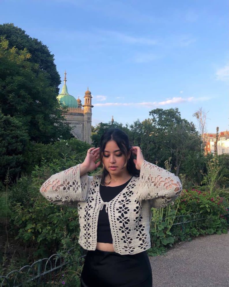 May include: A woman wearing a white crocheted cardigan with a tie front and bell sleeves. She is standing in front of a building with a green roof.