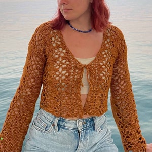 May include: A brown crochet cardigan with a tie front and bell sleeves. The cardigan is worn over a white tank top and blue jeans.