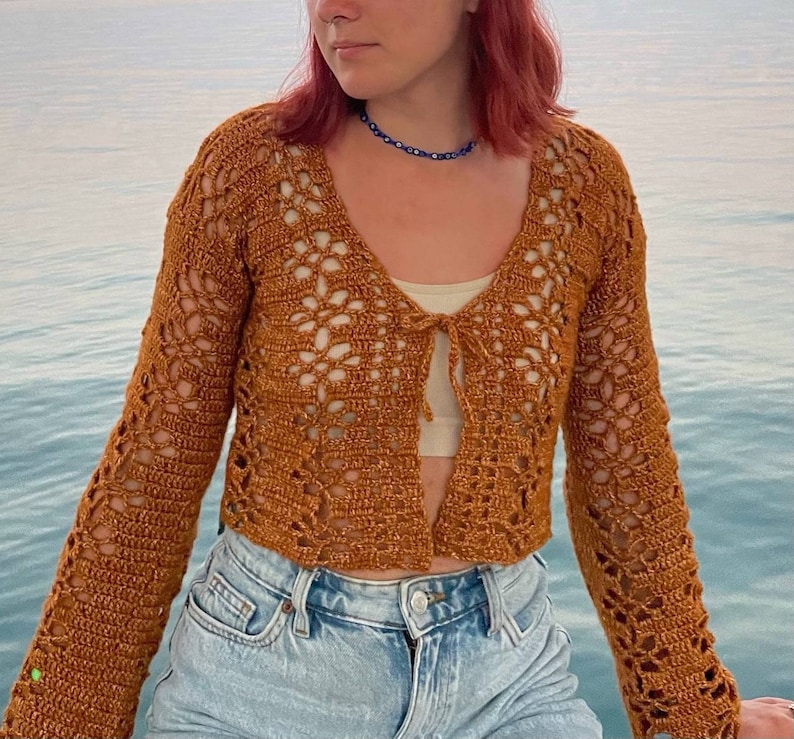 May include: A brown crochet cardigan with a tie front and bell sleeves. The cardigan is worn over a white tank top and blue jeans.