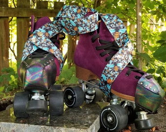 Puede incluir: Un par de patines de cuatro ruedas de ante morado oscuro con cordones negros, ruedas negras y punteras iridiscentes. Los patines están adornados con largas correas de tela decoradas con un patrón repetitivo de abejas amarillas y negras y pequeñas flores azules.