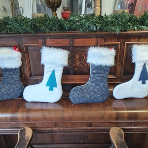 May include: Four white Christmas stockings with blue and white trim and a teal tree design. The stockings are hanging on a wooden piano.