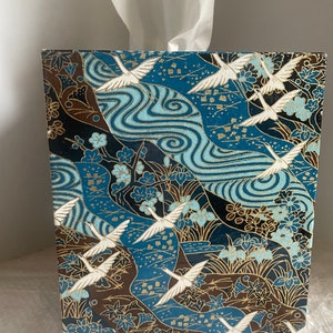 May include: Tissue box with a Japanese-inspired design. The box features a pattern of white cranes in flight over a blue river with gold accents. The background includes brown and navy blue floral and leaf patterns.