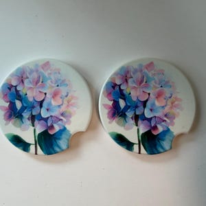 May include: Two circular car cup holder coasters featuring a white background with a vibrant hydrangea flower print in shades of blue, purple, and pink. Each coaster has a small semi-circular notch on the side for easy removal from the cup holder.
