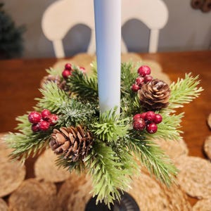 May include: A festive candle holder featuring a white candle surrounded by a wreath of green pine needles, red berries, and brown pine cones. The candle holder is designed for the holiday season and adds a touch of nature to any table setting.