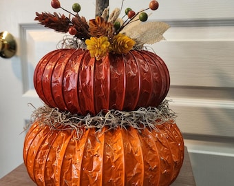 May include: A decorative two-tiered pumpkin with a rustic design. The top pumpkin is a deep red, while the bottom is orange. The pumpkins are adorned with dried foliage, berries, and a wooden stem, perfect for fall decor.