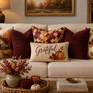 Pode incluir: Uma coleção de almofadas decorativas num sofá creme, incluindo uma almofada retangular com o texto Grateful e um gráfico de abóbora, ao lado de almofadas quadradas com cenas de floresta de outono, padrões xadrez e folhas de ácer.