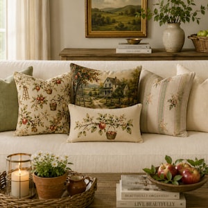 May include: A collection of decorative throw pillows on a sofa, featuring a solid sage green pillow, an apple-patterned square pillow, a landscape scene pillow, a rectangular apple-themed pillow, a striped floral pillow, and a solid cream pillow.