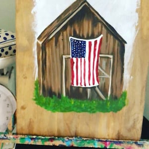 May include: A painting of a wooden barn with an American flag hanging from the side. The barn is painted brown and the flag is red, white, and blue. The barn is set against a white background.