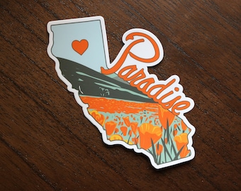 Op de afbeelding: Een sticker met een omtrek van de staat Californië met het woord "Paradise" in een gestileerd lettertype. De sticker heeft een blauwe en groene achtergrond met oranje bloemen en een rood hart in de linkerbovenhoek.