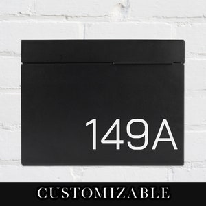 May include: Black metal mailbox with a white number "149A" on the front. The mailbox is mounted on a white brick wall.  The text "CUSTOMIZABLE" is printed below the mailbox.