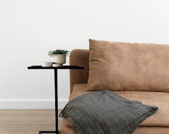 Peut inclure: Une table d'appoint en métal noir avec une base carrée et un plateau rectangulaire. La table est à côté d'un canapé en cuir marron avec une couverture en plaid gris et jaune.